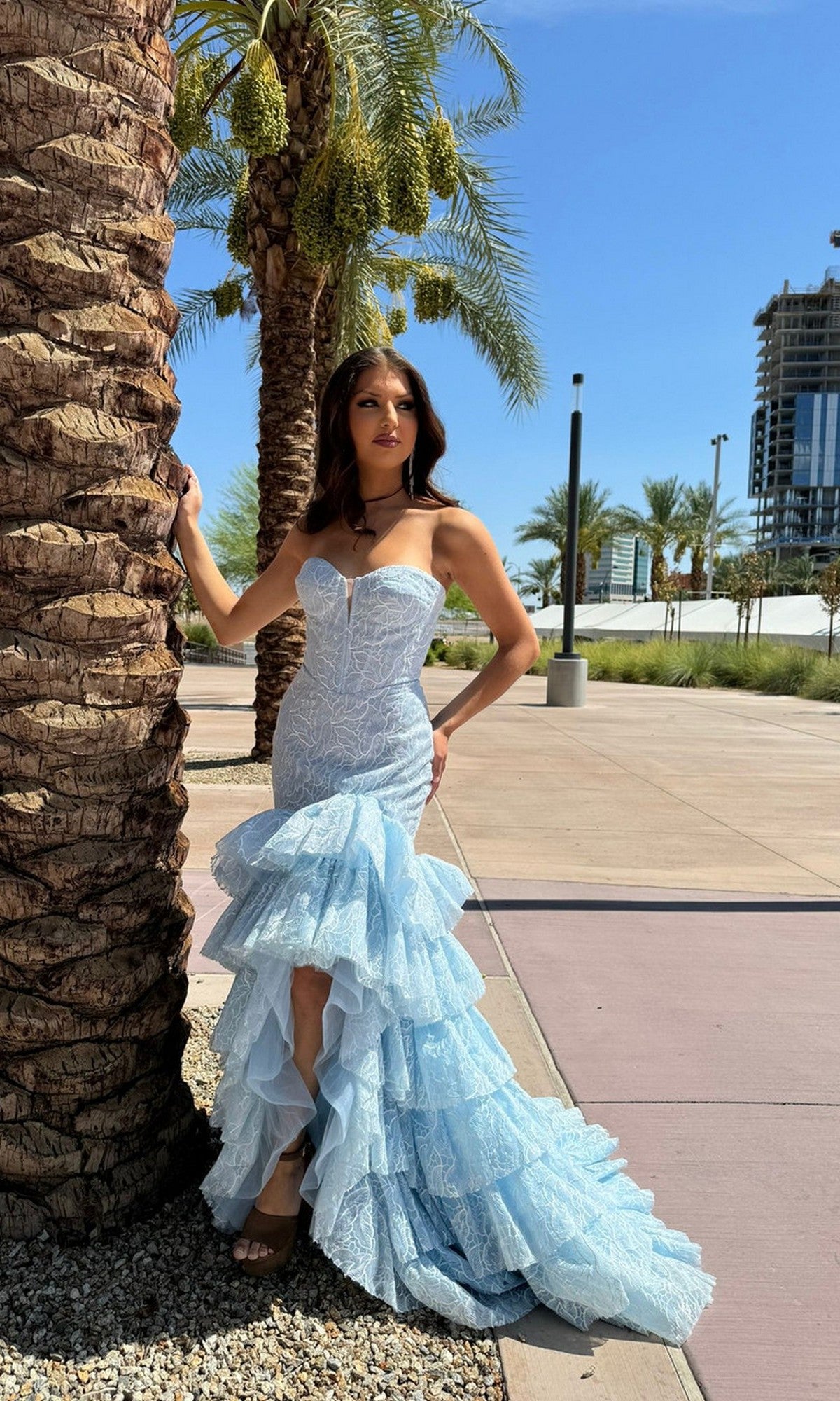 A woman wearing the Portia and Scarlett PS25250 long formal dress in blue, featuring a strapless boned corset bodice and ruffled tiered skirt, stands by a palm tree on a sunny day with modern buildings under a clear blue sky.